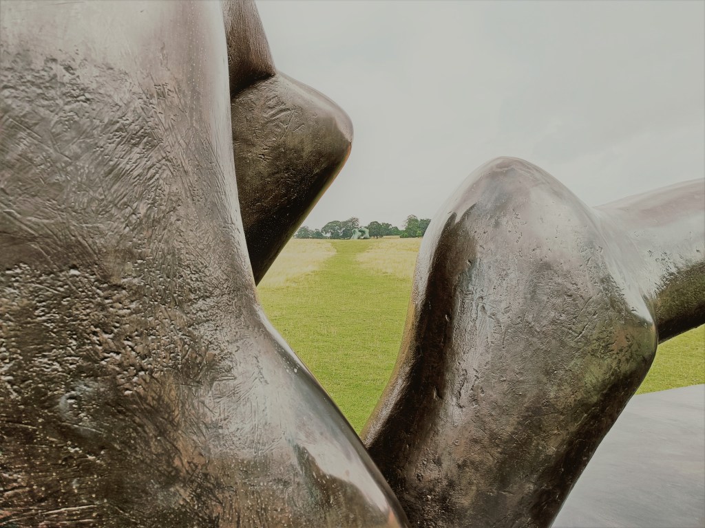 Detail of Henry Moore's sculpture showing curves of the figure and the texture of the bronze, with other exhibit in the far distance in the park.