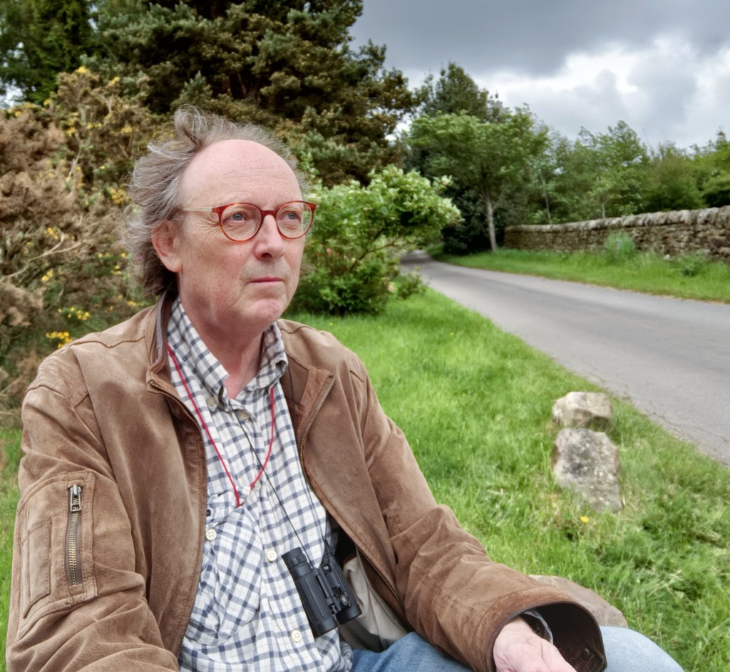Photograph of the composer seen sat on a rock by the side of a country lane. Dry stone wall, trees and dark clouds are in the background.