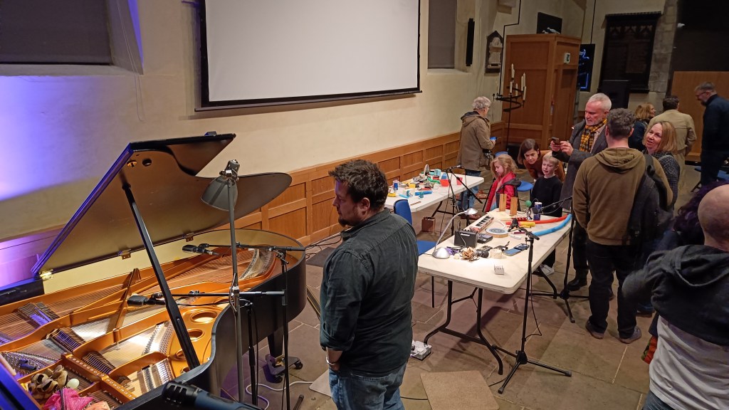 Members of the audience of all ages inspect the stage area: A grand piano left with lid open, to the right two tables full of the objects used to create the soundscape each table having microphone on a stand near by.