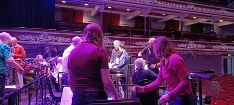 Musicians are setting up, the narrator and conductor are discussing the final arrangement before the performance in the ornate theatre hall