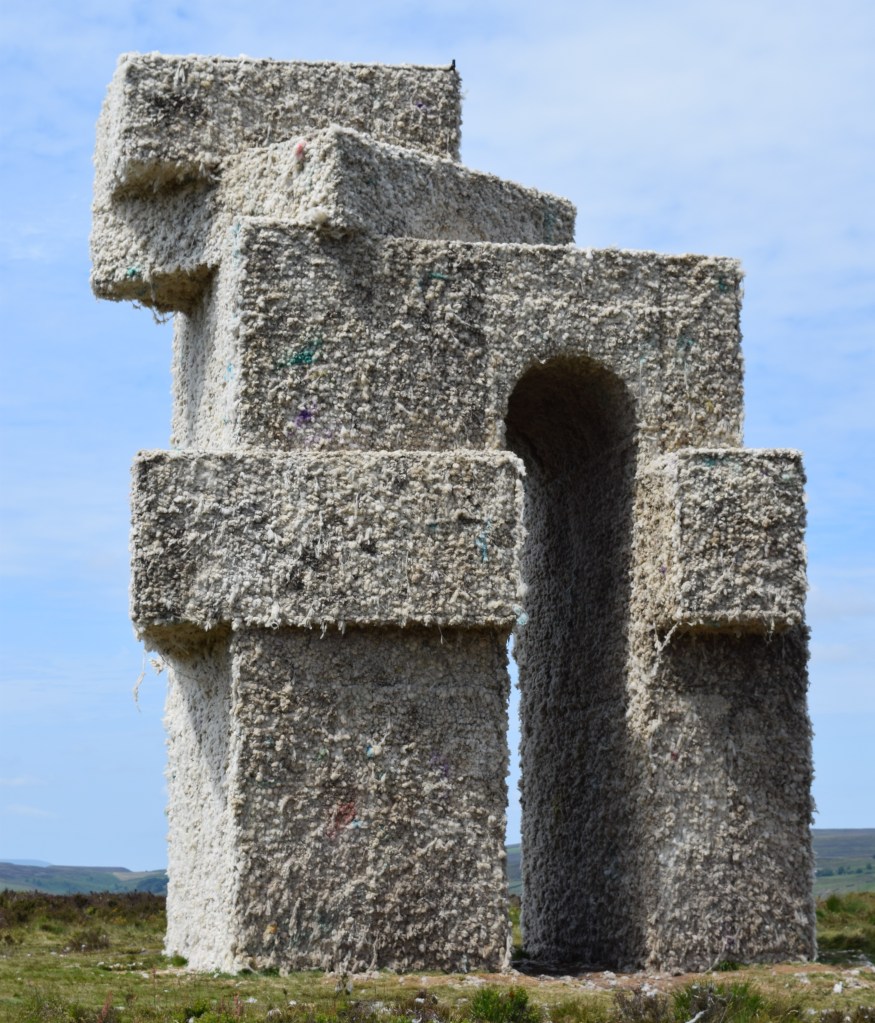 A large grey white rectangular structure looking like a series of boxes stacked at different angle there is a tall arch going through the middle of the structure.