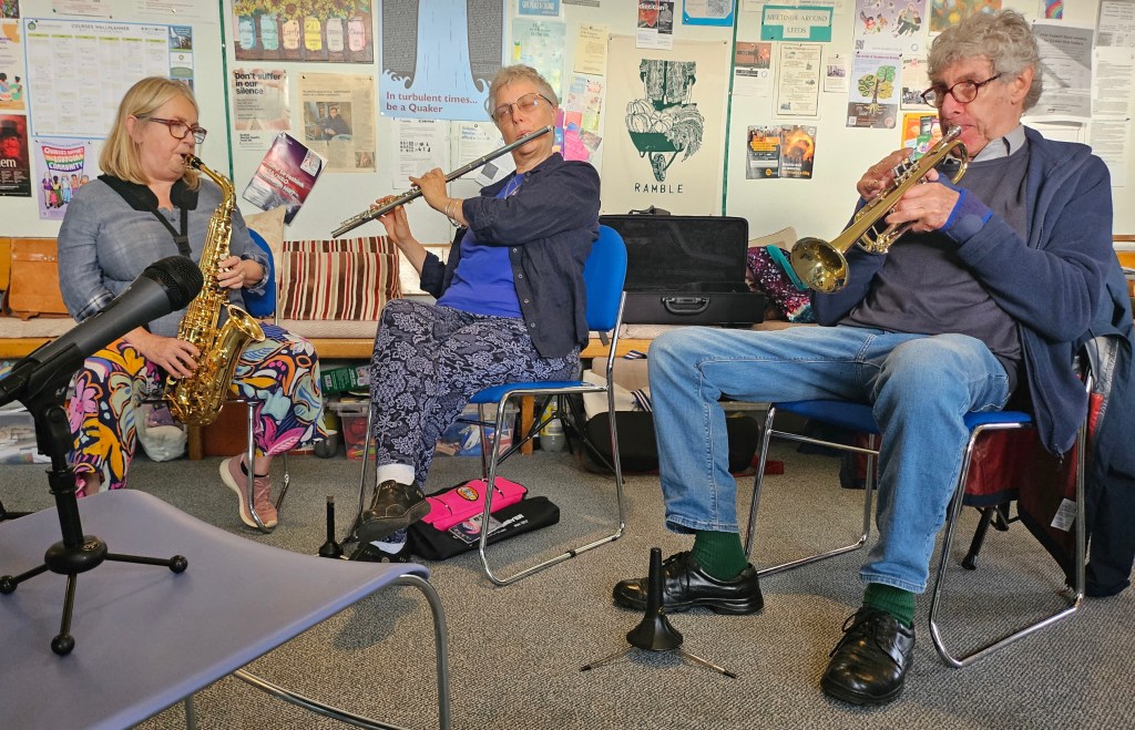Microphone foreground with musician playing left to right: Alto Saxophone, Flute, and Trumpet.