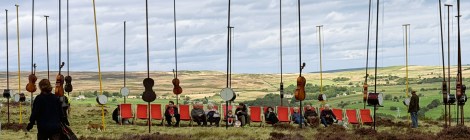 Moorland heather and grass landscape, with an array of 4 metre vertical poles some with cello bodies other with drums attached to act has resonators for the harp string mounted lengthways across each pole. A row deck chairs are in the middle of the installation with people sat down listening.