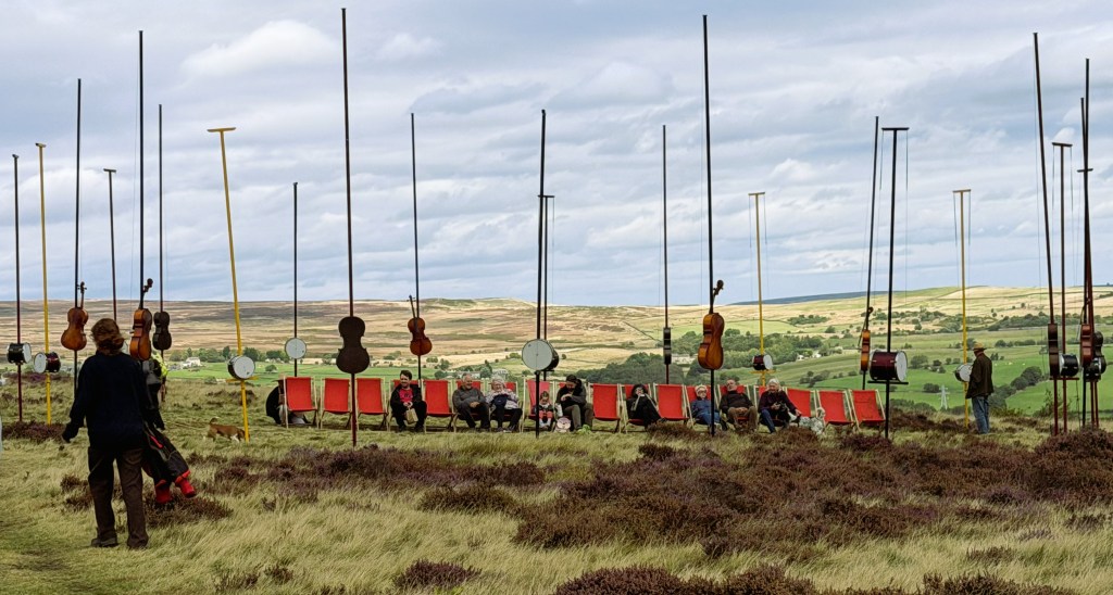 Moorland heather and grass landscape, with an array of 4 metre vertical poles some with cello bodies other with drums attached to act has resonators for the harp string mounted lengthways across each pole. A row deck chairs are in the middle of the installation with people sat down listening.
